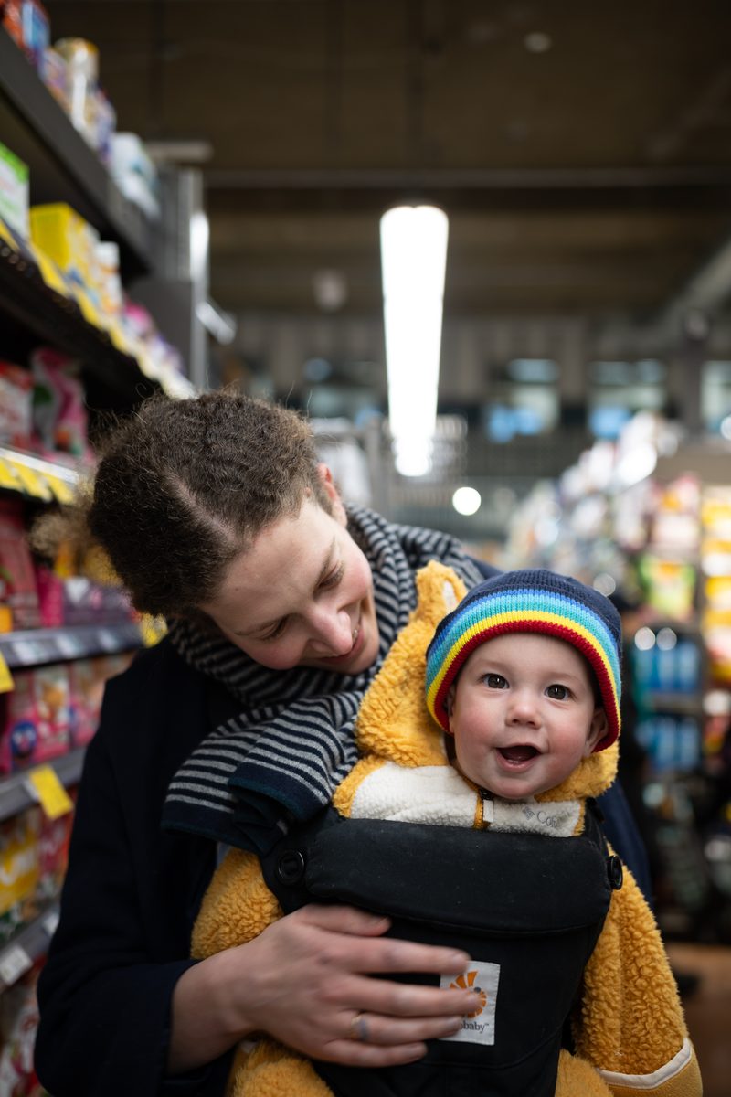 A mother smiles down at her baby, who wears a rainbow knit hat in a front carrier, in an aisle of the Whole Foods supermarket on South Street.