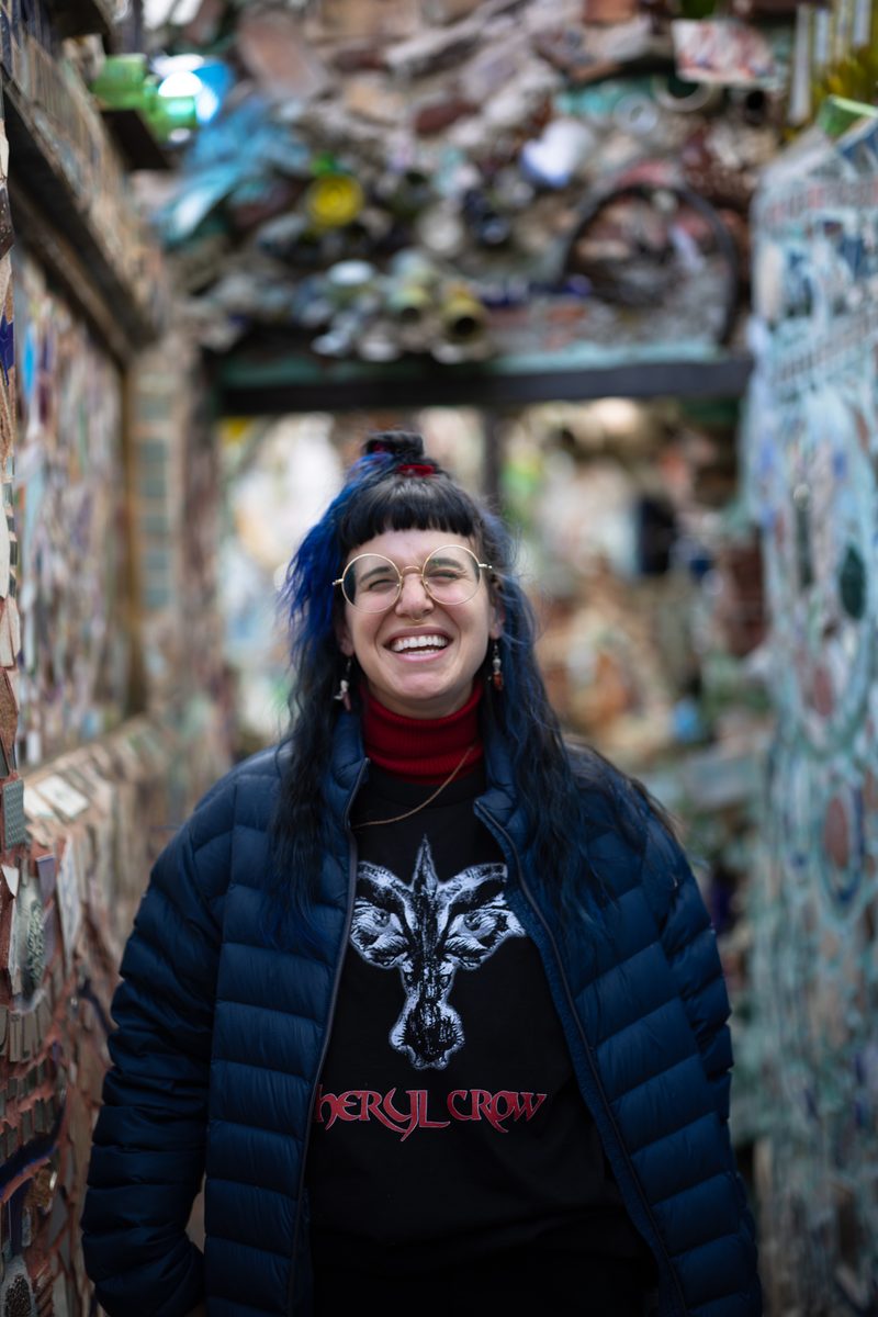 A woman with blue-tipped hair and glasses laughs in front of the mosaic walls of Philadelphia's Magic Gardens.