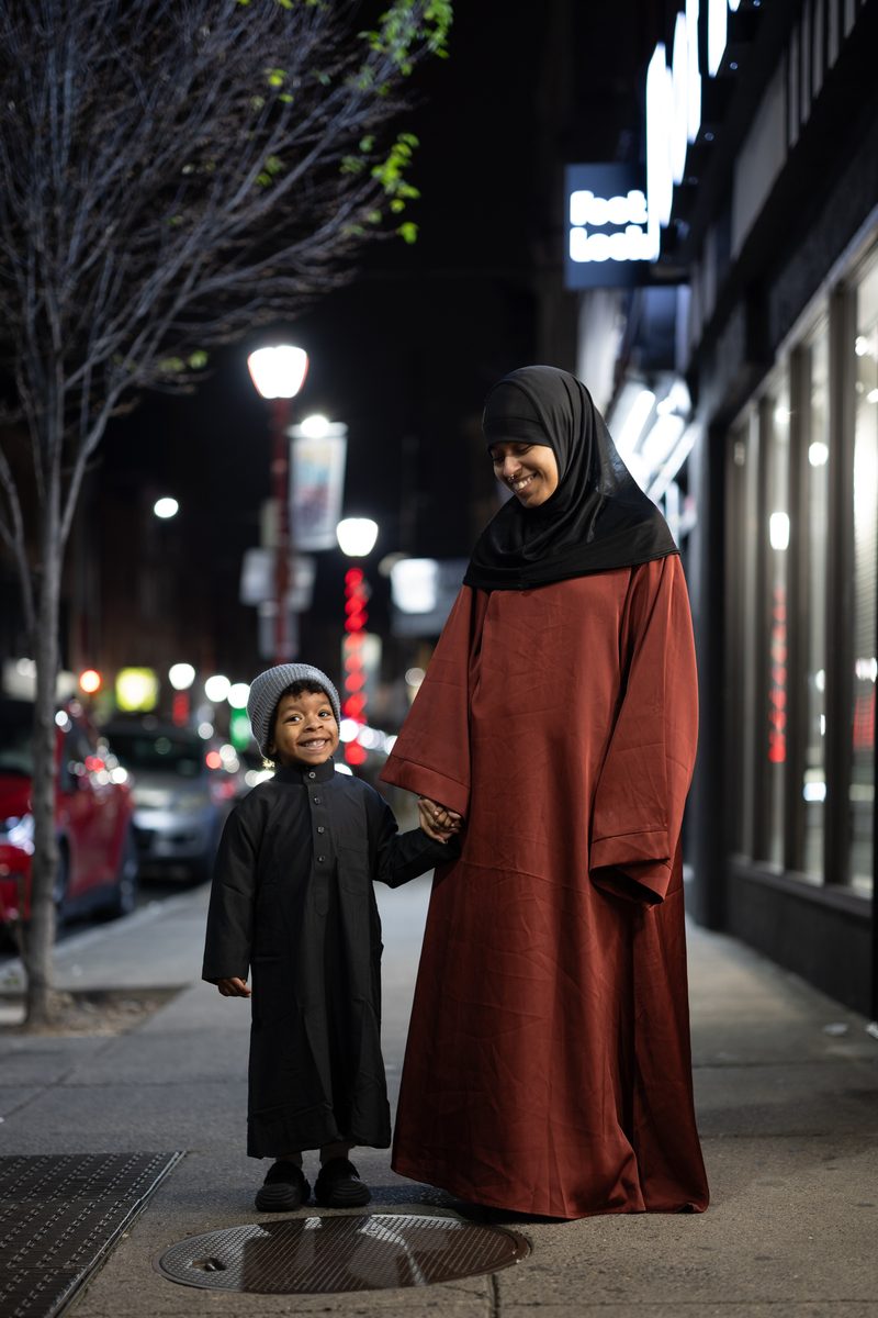 A woman in a hijab and rust-colored dress holds hands with a young boy in a dark robe on South Street at night.