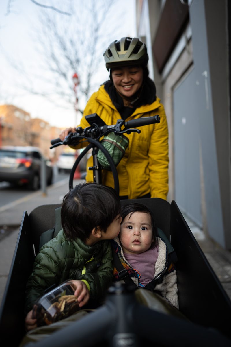 A mother in a yellow jacket and bike helmet rides a cargo bike with two young children in the front box.