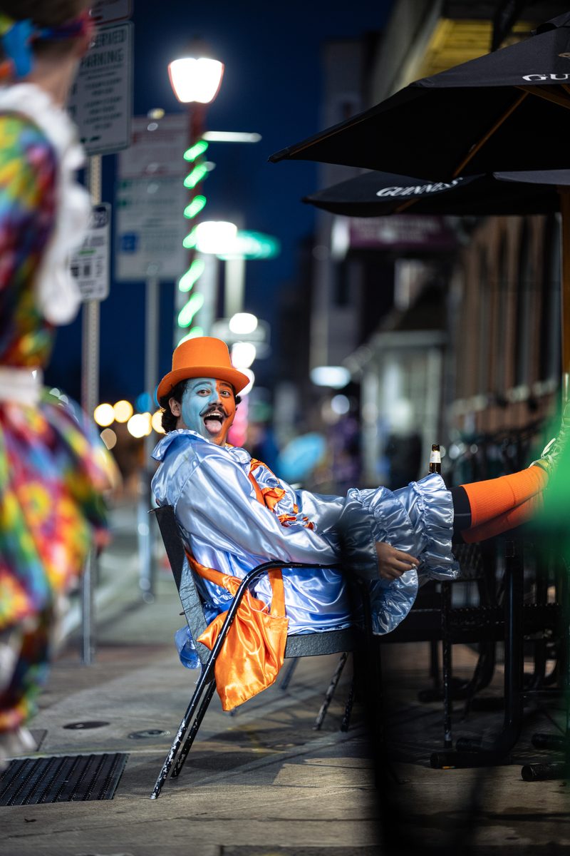 A Mummer in blue satin, an orange top hat, and painted face laughs at a sidewalk table on 2 Street after the New Year's Day parade.
