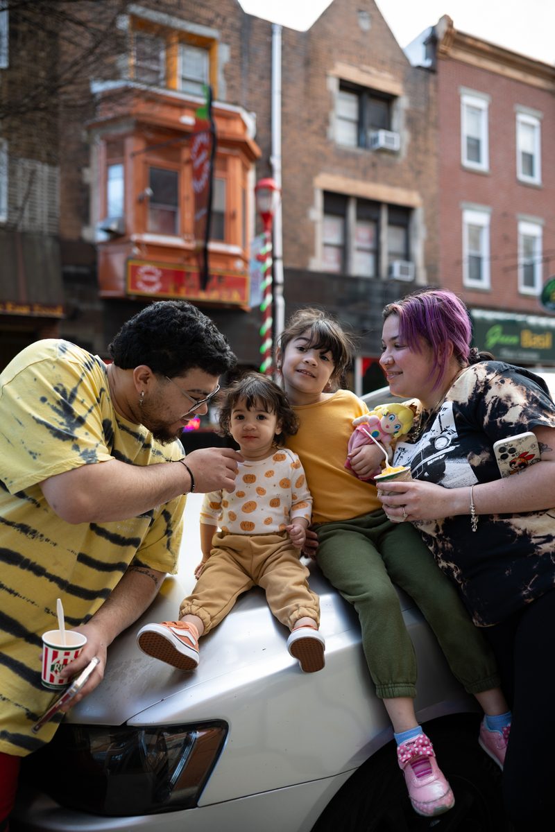 A family of four poses on the hood of a parked car, with the father feeding his toddler daughter a spoonful of ice cream.