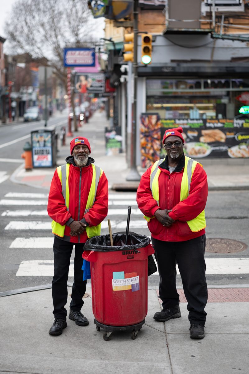 Two sanitation workers in red caps and reflective vests stand with their cart at a South Street crosswalk.