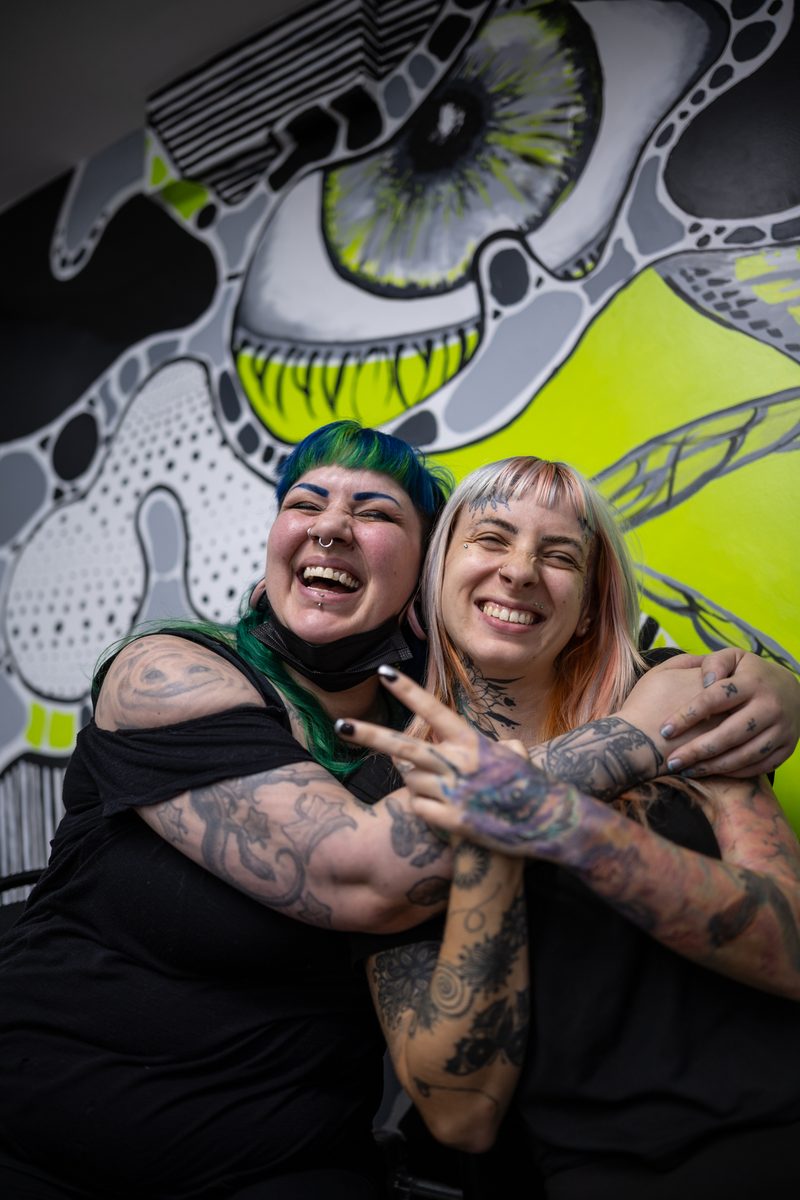 Two tattooed friends embrace and laugh in front of a black, white, and neon-green eye mural.