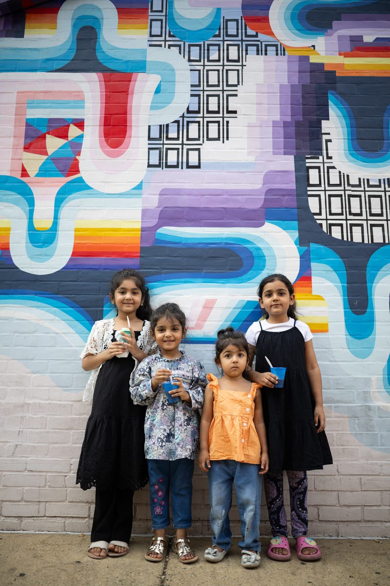 Four young children in dresses and sandals line up in front of a colorful geometric mural.