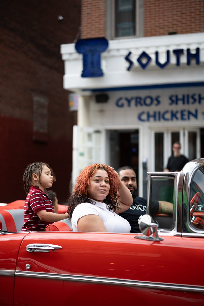 A couple rides in a red vintage convertible past Greek restaurant South Street Souvlaki.