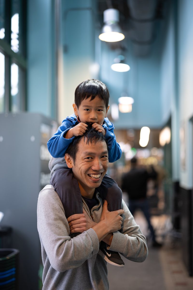 A father carries his young son on his shoulders inside a Whole Foods supermarket on South Street. Both are smiling.