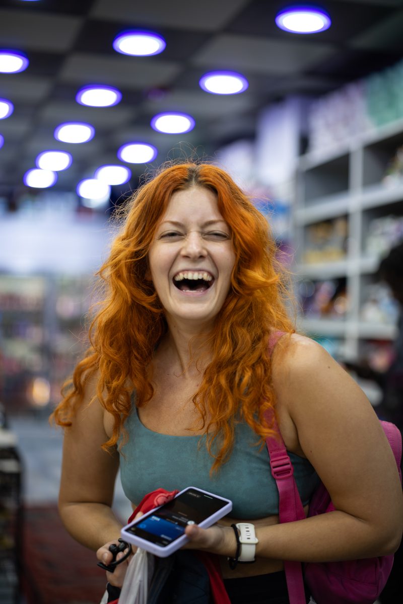 A woman with long red hair laughs with her phone in hand inside a brightly lit store.