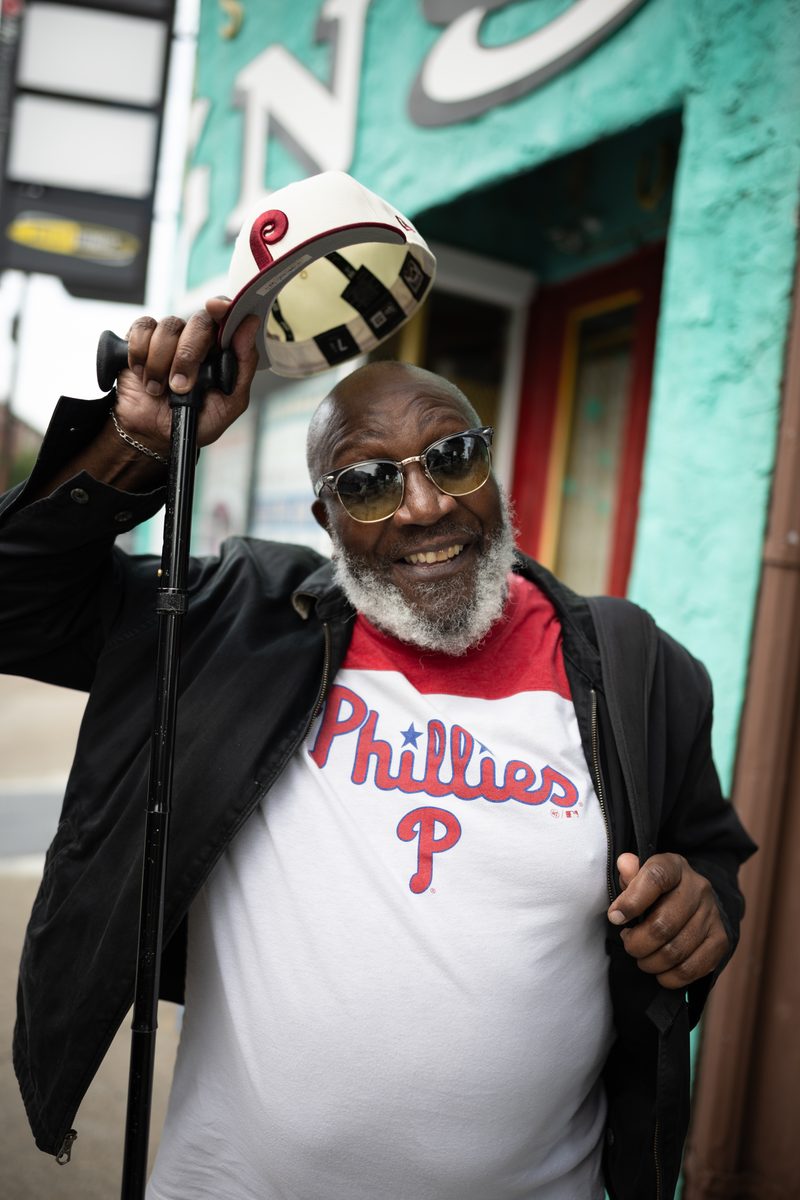 A man with a gray beard and gold sunglasses tips his Phillies cap, leaning on a cane in a Phillies t-shirt.