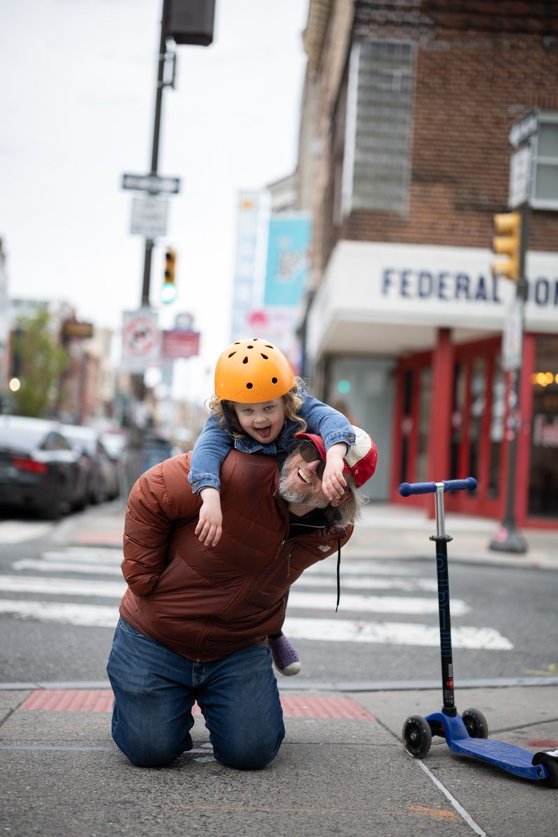 A parent kneels at a South Street crosswalk with a laughing toddler in an orange helmet on their back, scooter beside them.