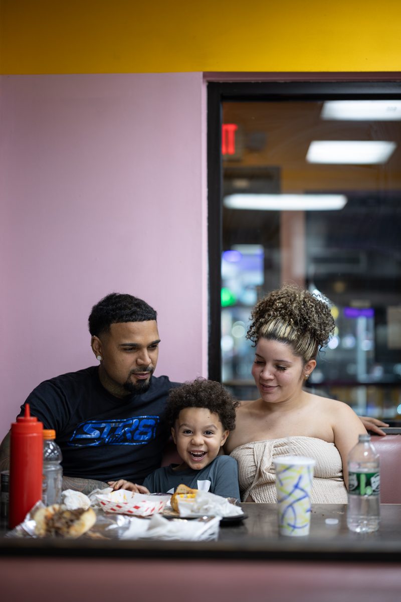 A young family of three shares a meal in a corner booth at Philly cheesesteak restaurant Ishkabibble's, with the child grinning between the parents.