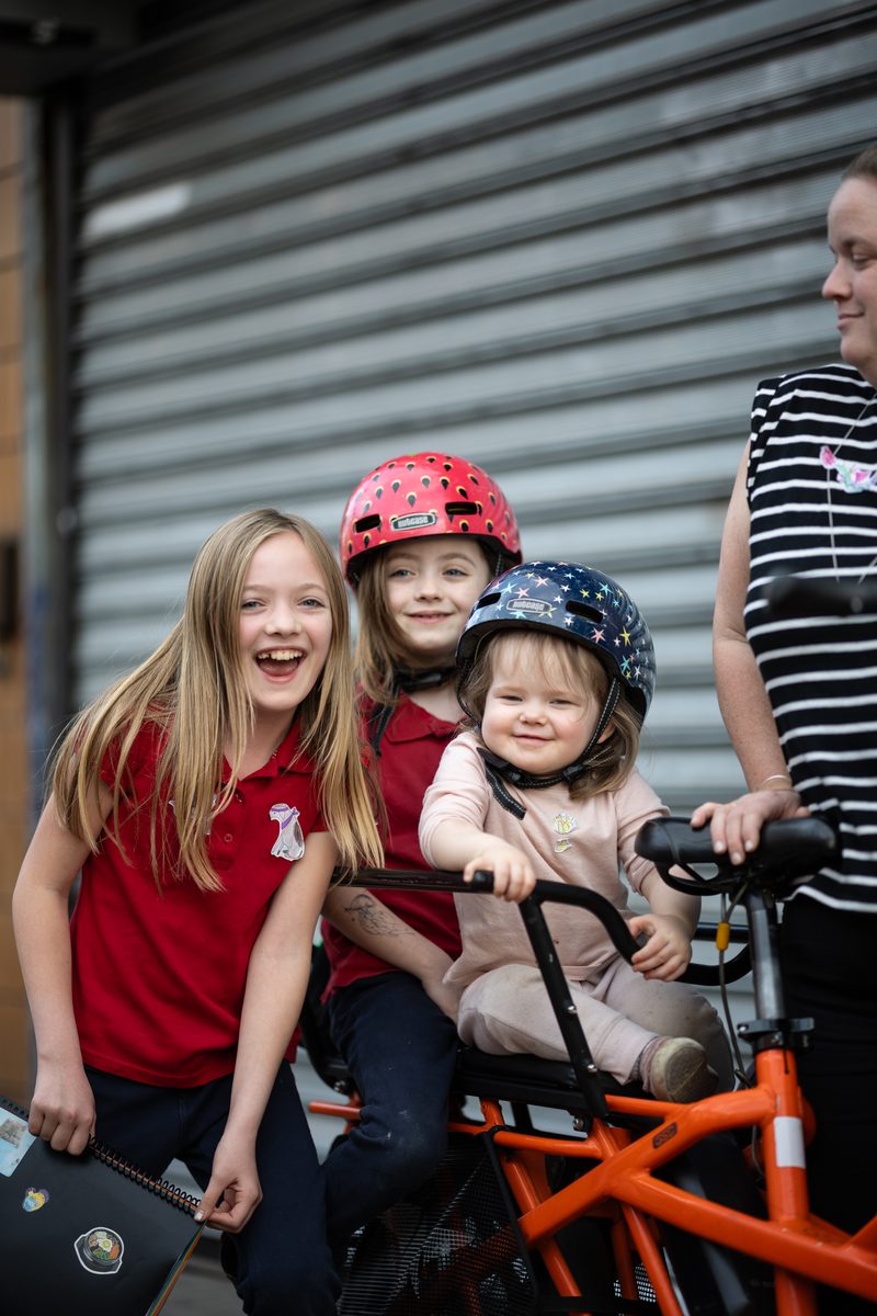 Three young sisters in bike helmets giggle on the front of a cargo bike, the smallest holding the handlebar.