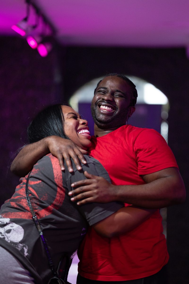 A couple laughs mid-embrace under pink and purple lights.