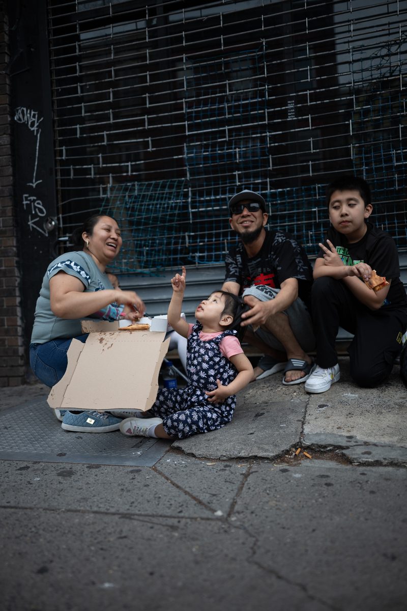 A family shares a pizza on the sidewalk in front of a shuttered storefront, the toddler pointing up at the sky during a 2024 solar eclipse in Philadelphia.