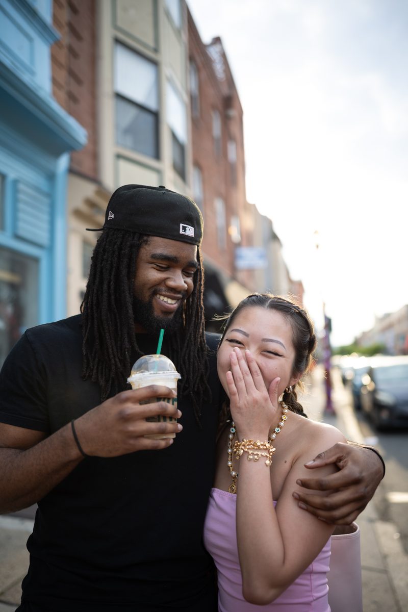 A man with locs and a black cap holds a Starbucks iced coffee with his arm around a laughing woman in pink.