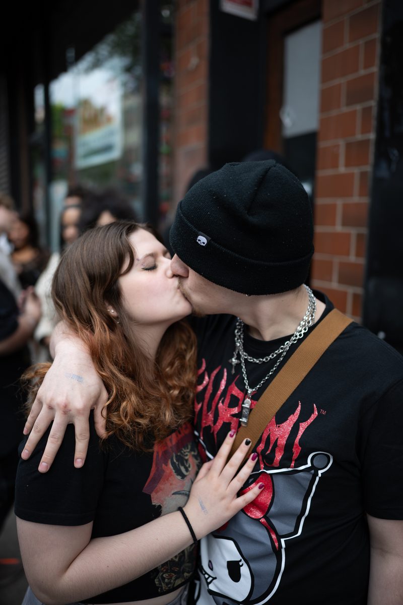 A young couple kisses on the sidewalk, she in a black band t-shirt, he in a black beanie.