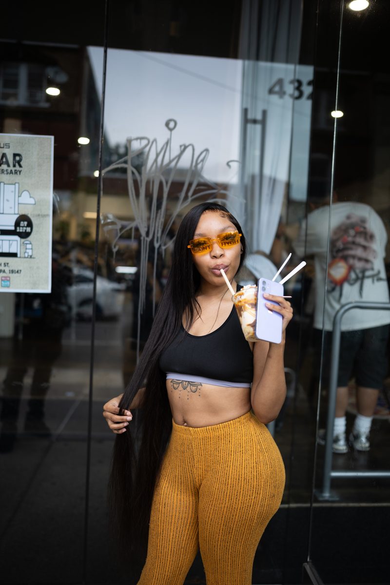 A woman in orange sunglasses and mustard pants sips a drink in front of Status shoe store while holding her long, waist-length hair with her right hand.