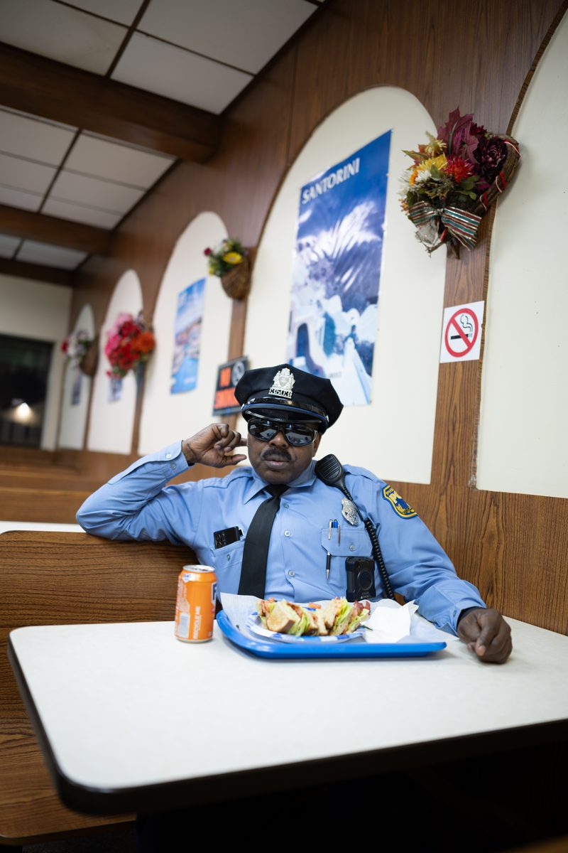 A Philadelphia police officer salutes from a booth at a South Street pizza shop, a tray of food with a Philly cheesesteak and an orange soda on the table.