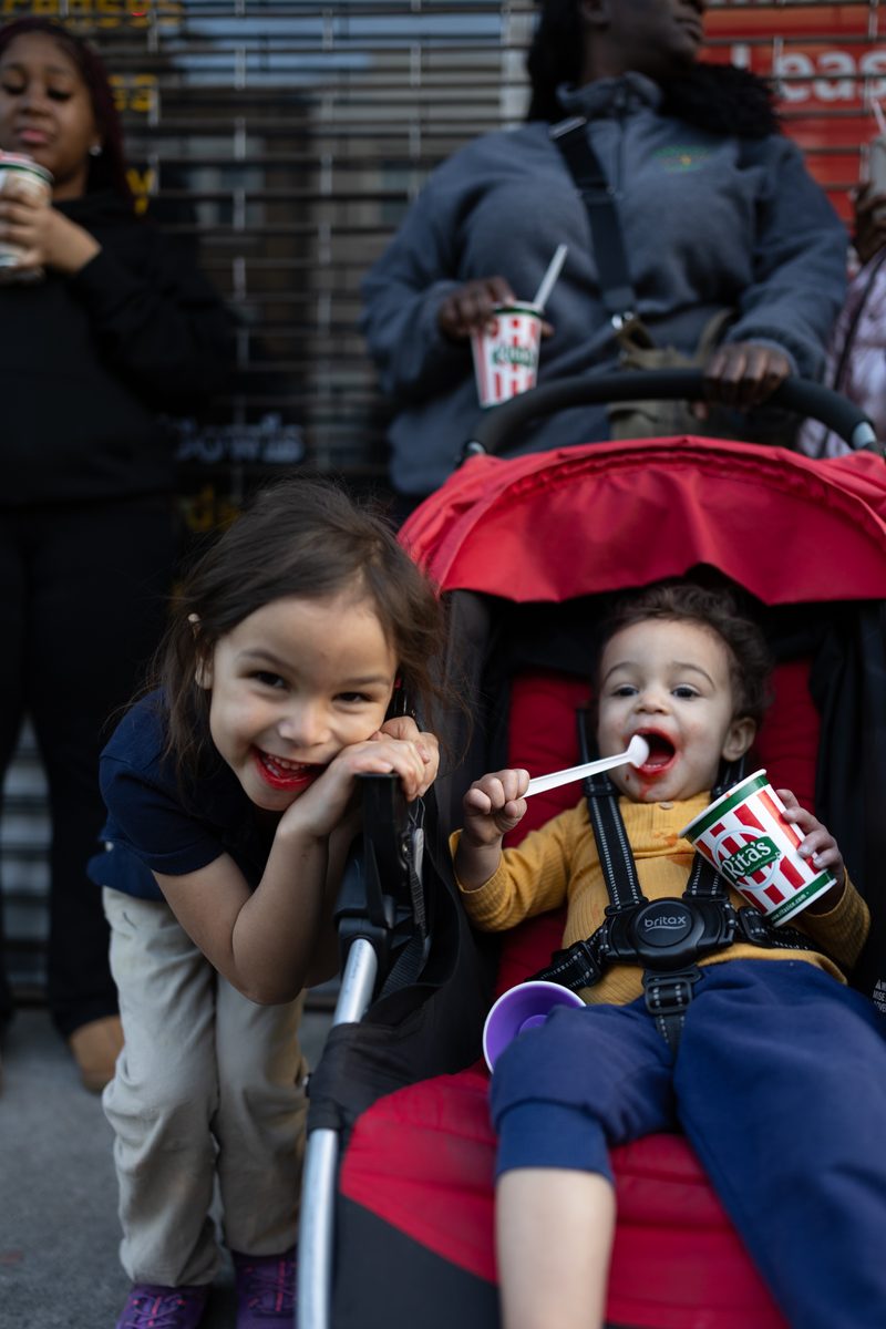 Two small children laugh in a red stroller on South Street, one eating a water ice with a spoon from a Rita's Water Ice cup.
