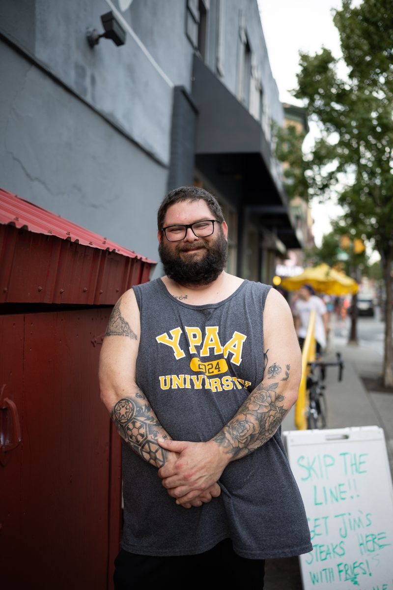 A bearded man with tattooed arms in a "YPAA University" tank top stands on the sidewalk outside Milkboy on South Street, and next to a Jim's Steaks sign.