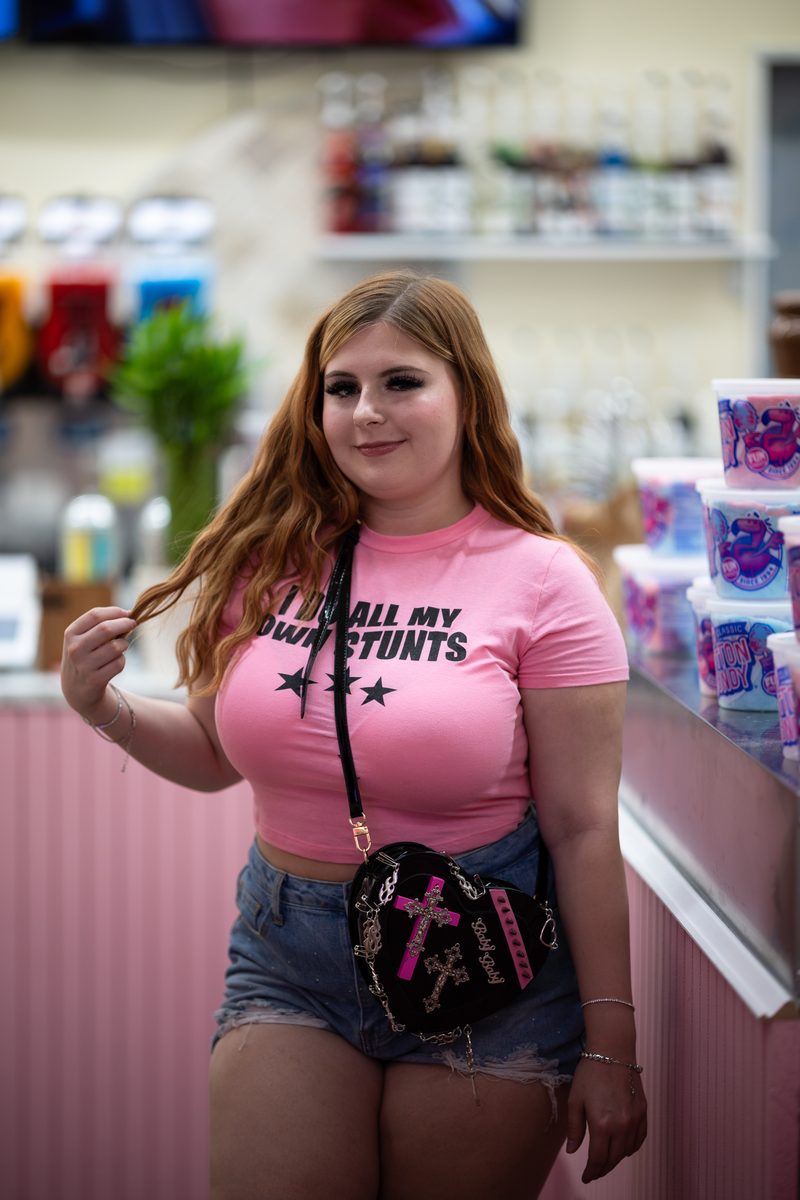 A young woman with long red hair in a pink "I Do All My Own Stunts" t-shirt poses inside a candy and ice cream store on South Street.