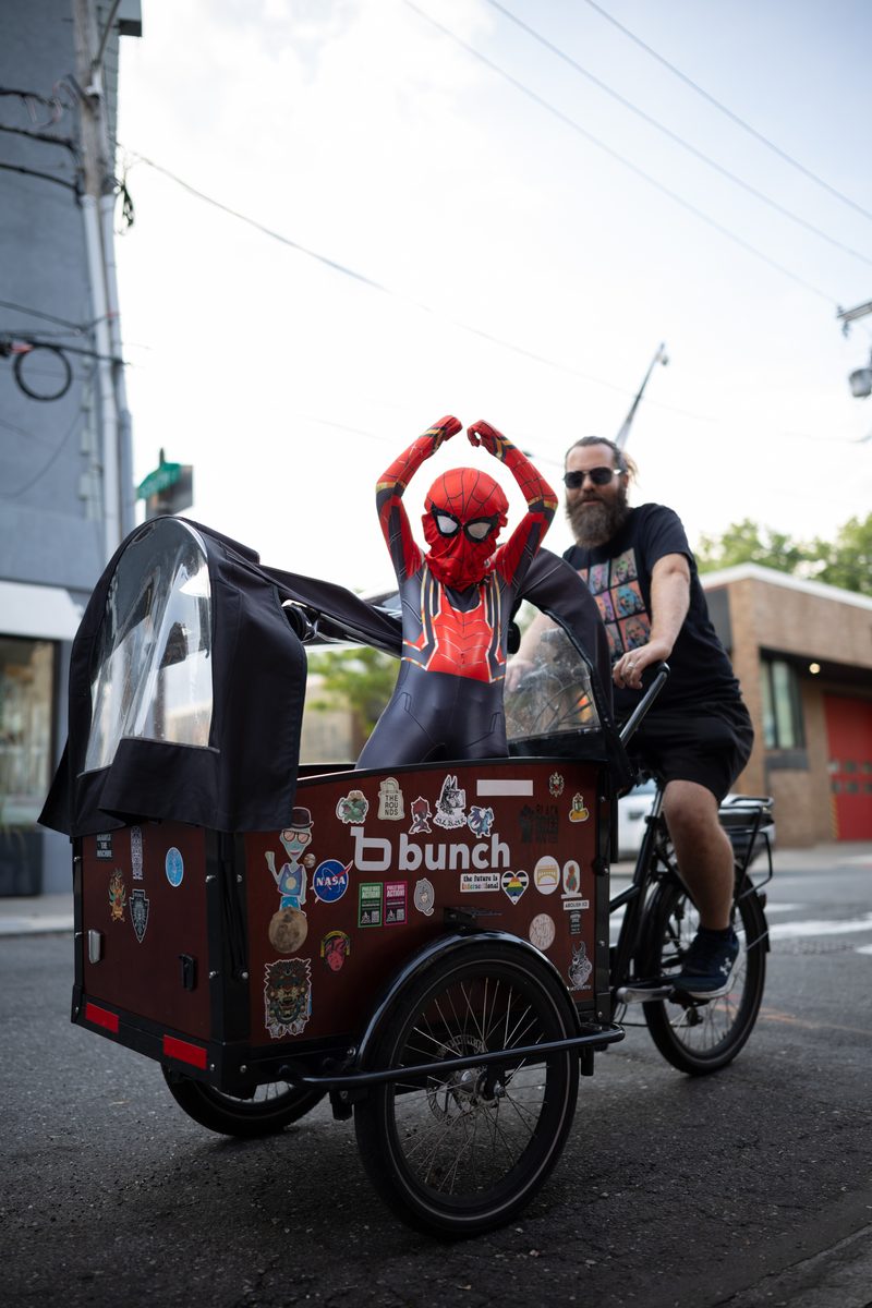 A child in a Spider-Man costume raises both arms from the front box of a cargo bike as their father pedals down the street.