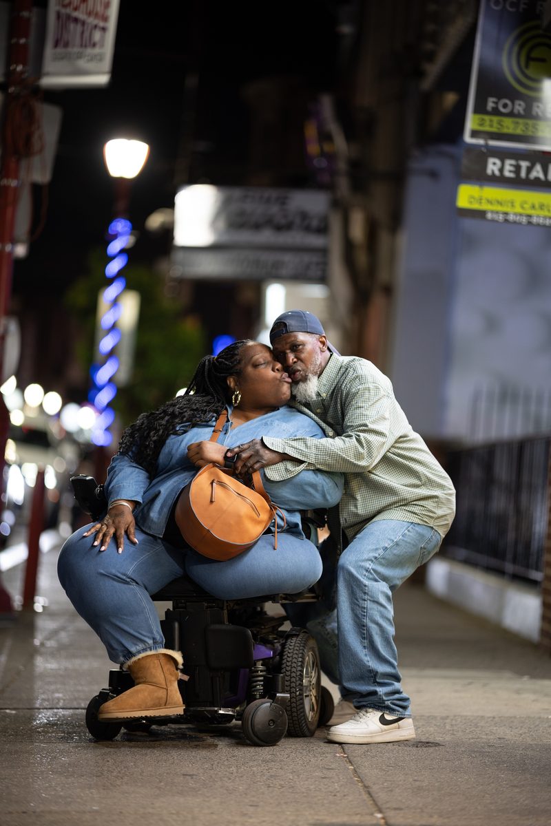 A man in a tan jacket kisses a woman seated in her power wheelchair on the sidewalk on South Street at night.