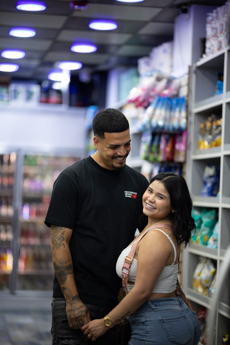 A young couple smiles together in the aisle of a convenience store, his arms around her from behind.
