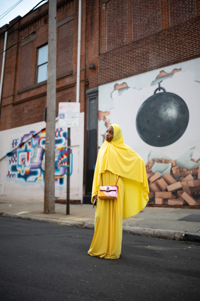 A woman in a bright yellow abaya and hijab with a pink handbag crosses the street in front of a wrecking-ball mural.