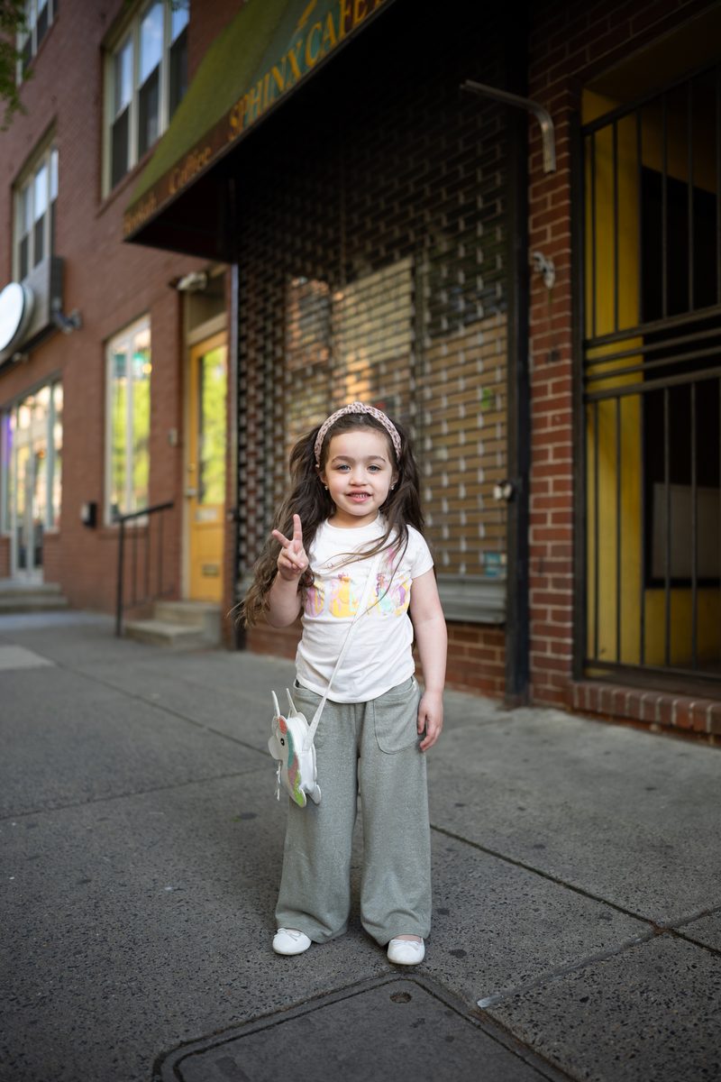 A little girl in a headband and retro green pants flashes a peace sign on the sidewalk outside a South Street storefront.