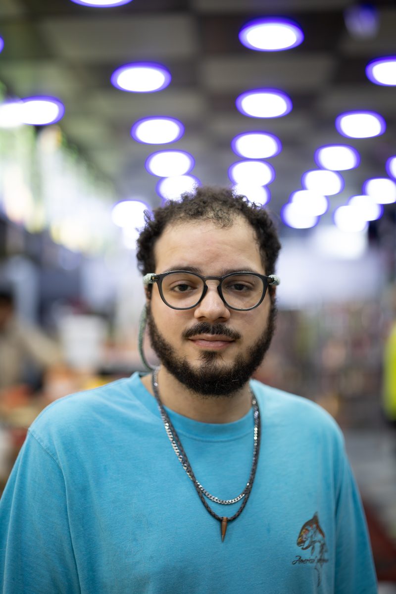 A man with glasses, a beard, and a blue t-shirt looks into the camera under the purple lights of a shop.