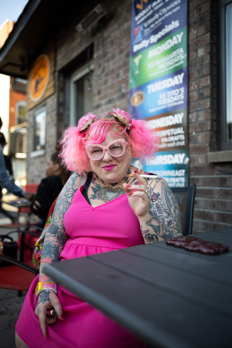 A woman with pink hair, pink glasses, and full-sleeve tattoos in a pink dress sits at an outdoor table.