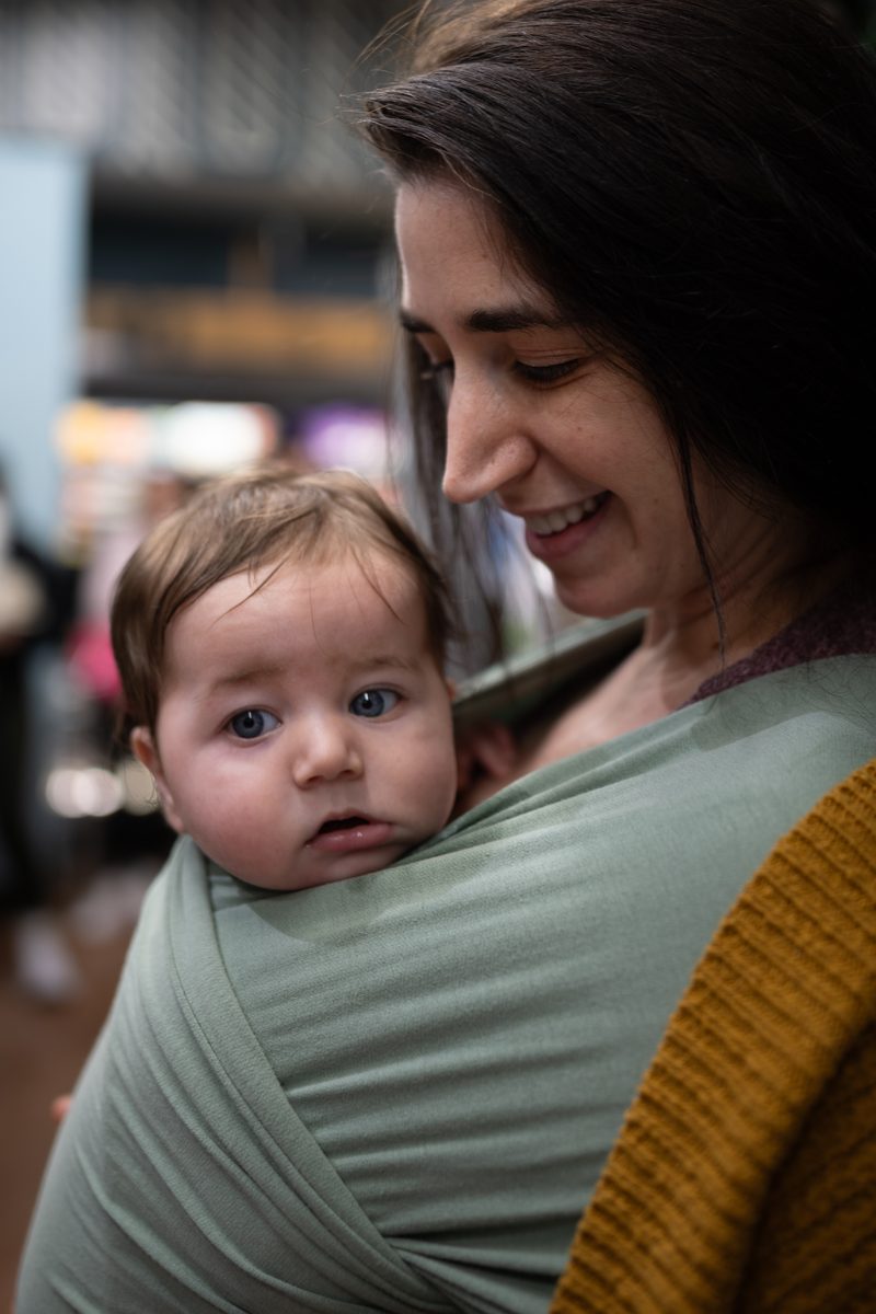 A mother smiles down at her baby, who looks at the camera from a sage-green wrap on her chest.