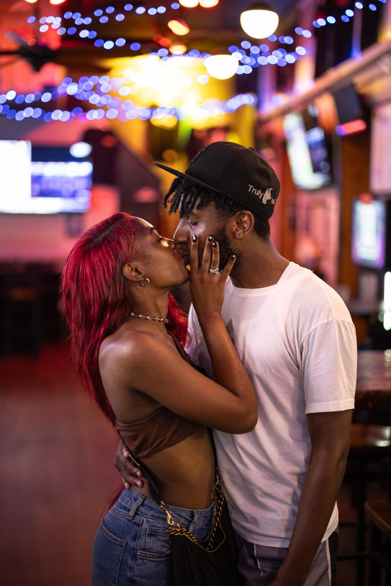 A couple kisses under string lights inside a South Street bar, she with red hair, he in a black cap.