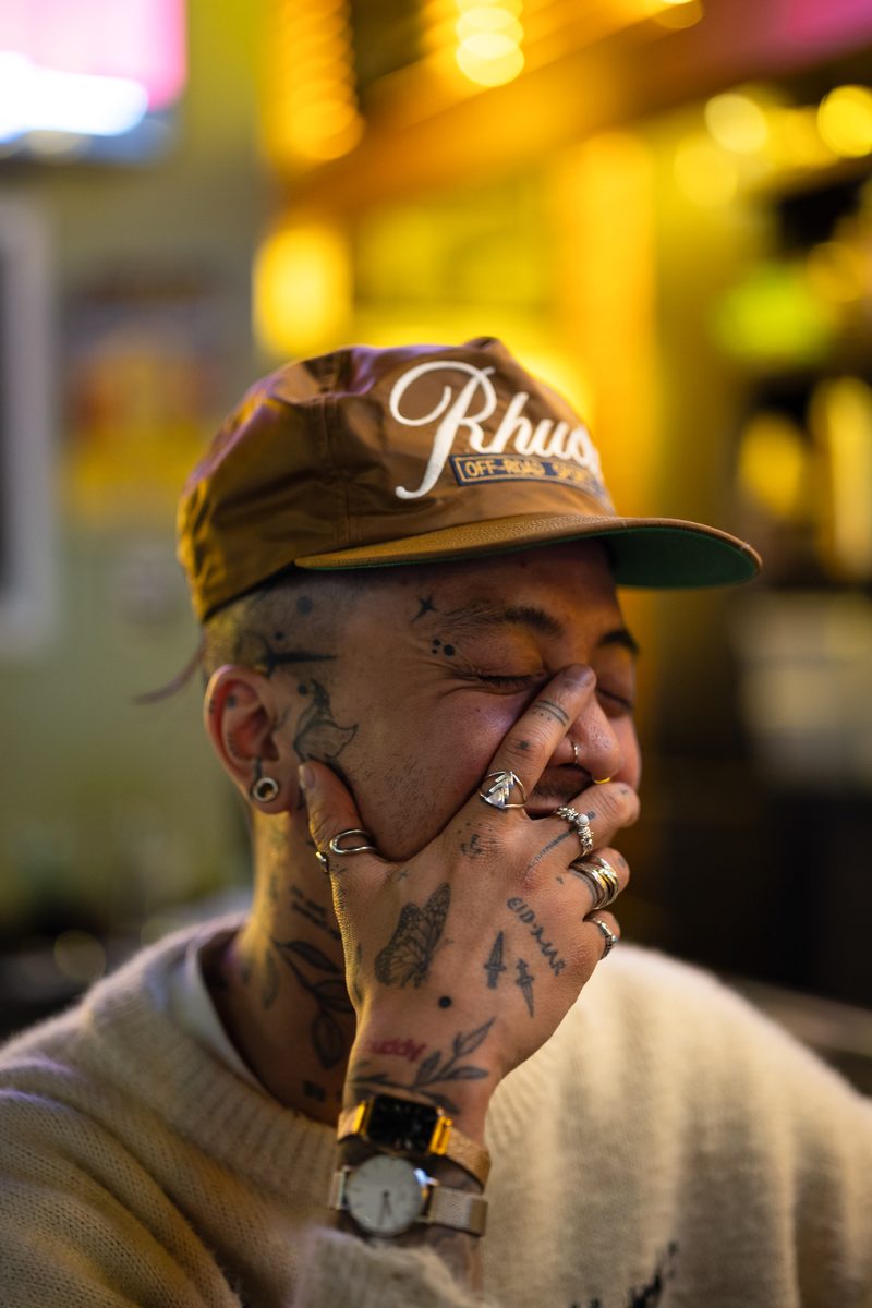 A man with face tattoos and a tan cap covers his face with a ringed hand, laughing.