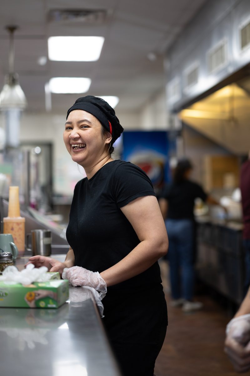 A woman in a black chef's cap and gloves smiles from behind the counter of South Street Philly cheesesteak restaurant Ishkabibble's kitchen.