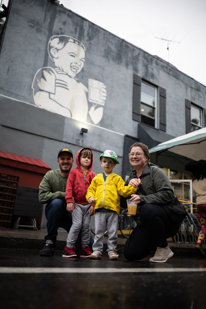A family of four sits on a curb beneath a mural of a smiling child holding a mug outside of Milkboy bar on South Street. The two kids are in red and yellow raincoats and the parents are in green.