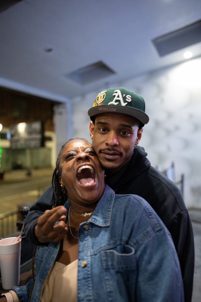 A laughing woman leans back into a man in a green A's cap and denim jacket, both holding drinks, at night at Fat Tuesday bar on South Street.