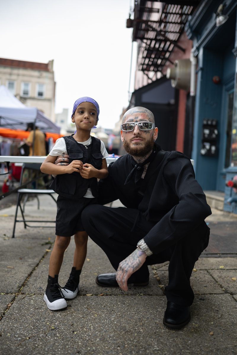 A tattooed man in all black crouches beside his young son in a purple headband on a South Street sidewalk.