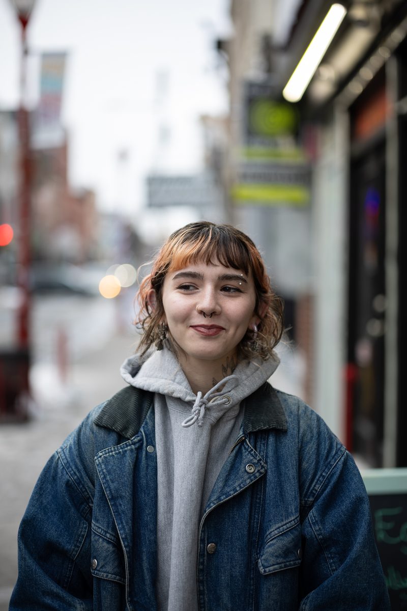 A young woman with copper bangs in an oversized denim jacket smiles softly on South Street.