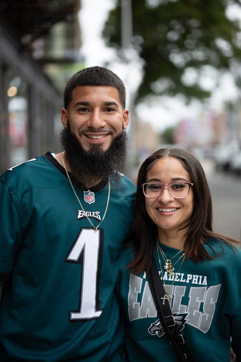 A couple in Philadelphia Eagles gear smiles on South Street, he in a number 1 Jalen Hurts jersey, she in a green Eagles sweatshirt.