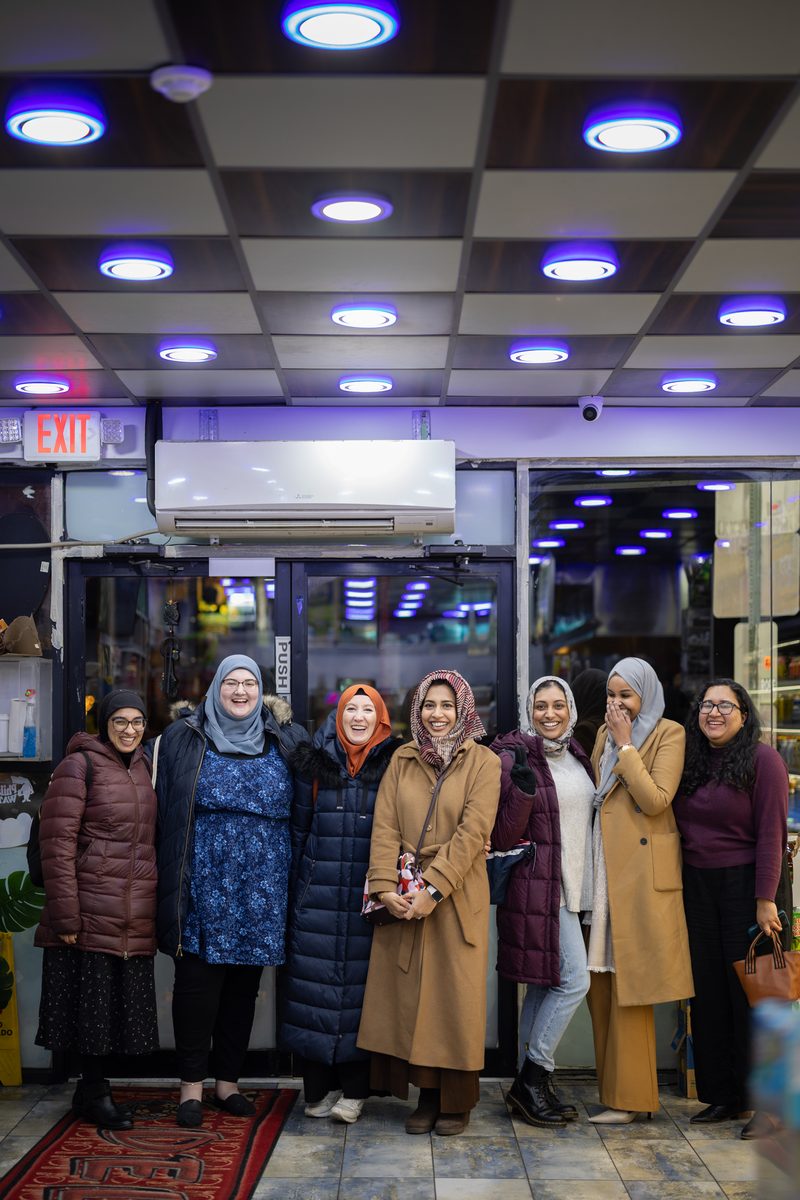 Seven women in hijabs and winter coats stand together smiling inside the entrance of a South Street corner store.