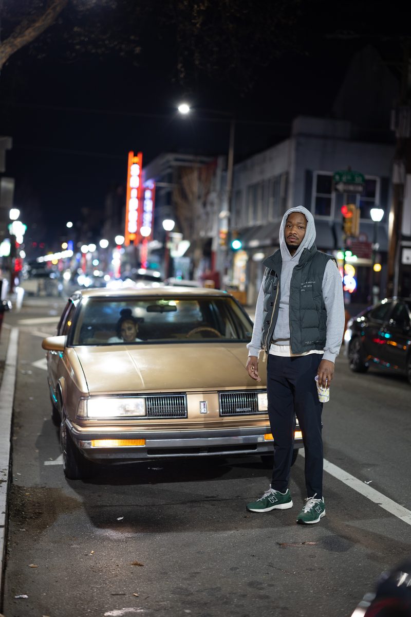 A man in a gray vest and hoodie stands in front of a gold vintage Pontiac sedan in the middle of South Street at night.