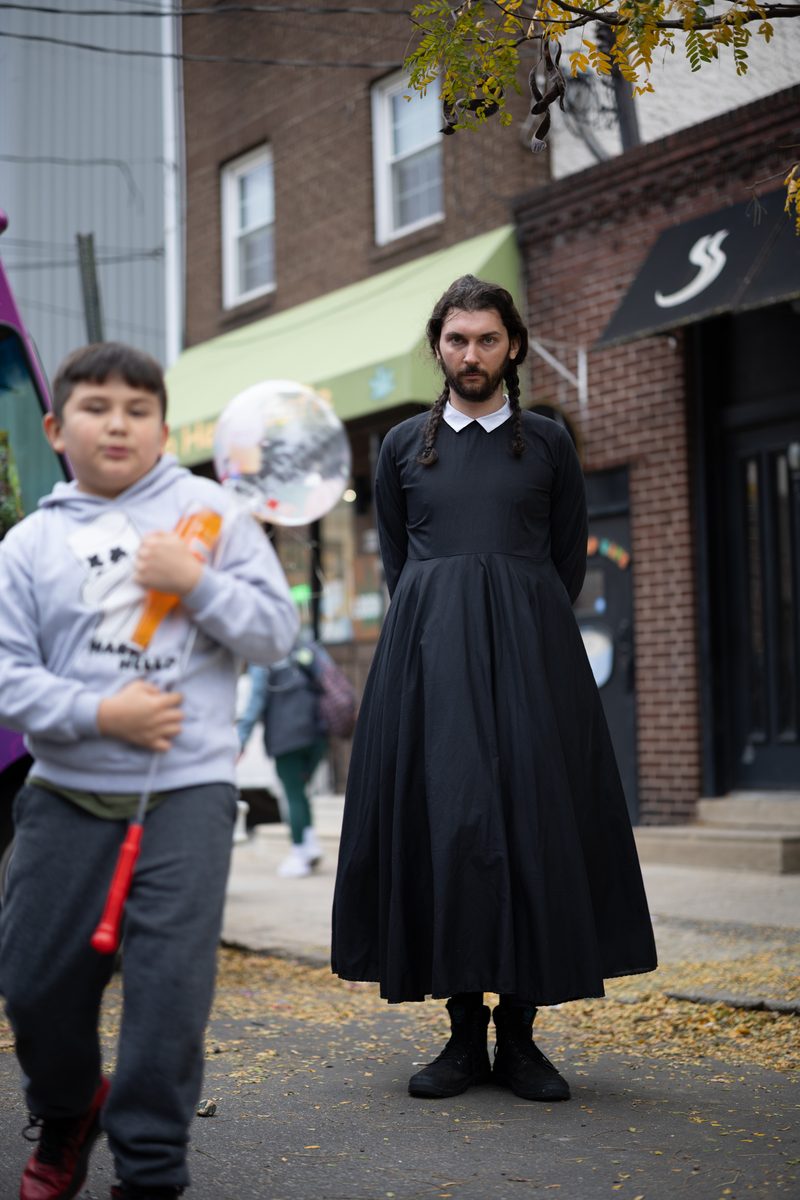 A man dressed as Wednesday Addams in a long black dress and braids stands on a South Street sidewalk on Halloween, a boy with a balloon beside him.