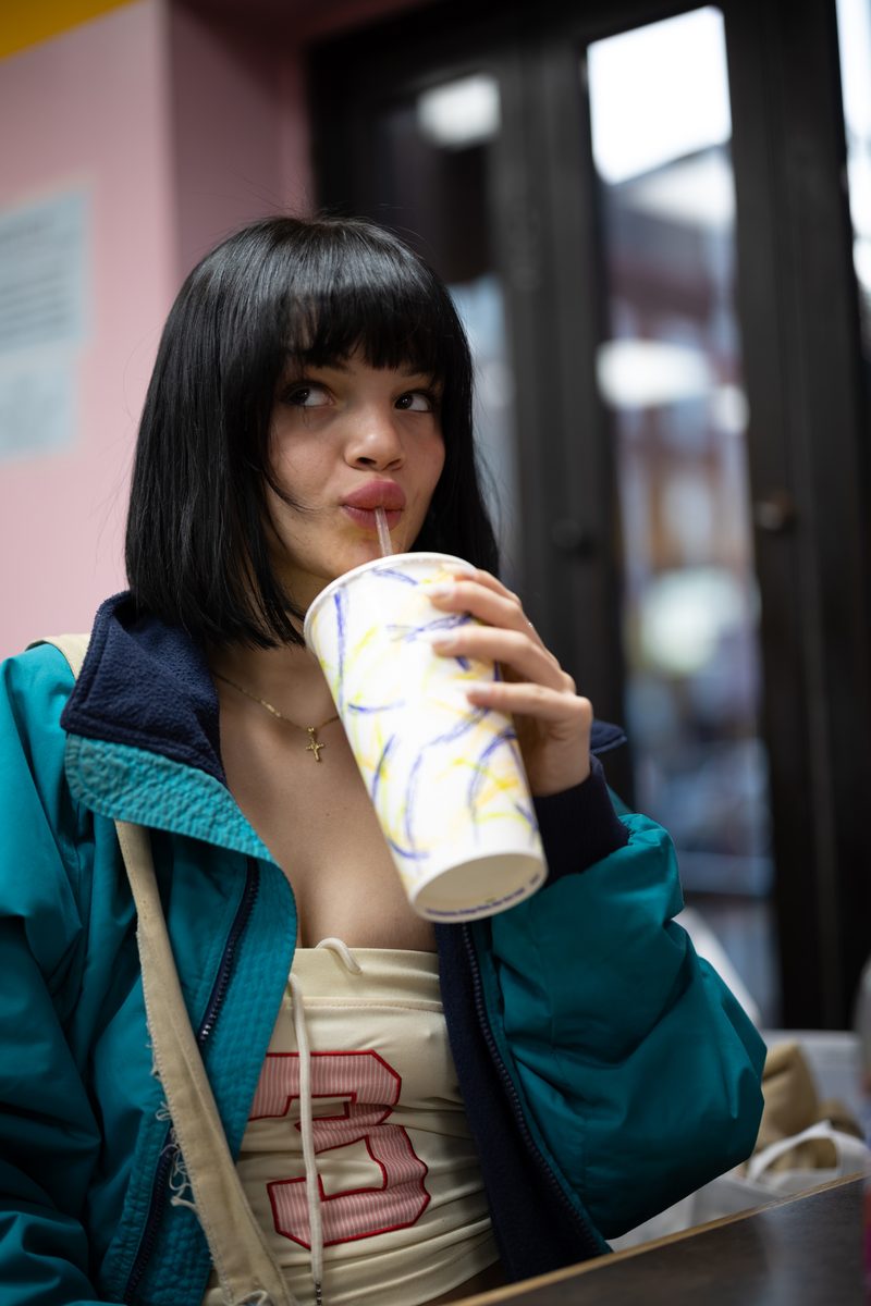A woman with a black bob and bangs sips a drink through a straw while side-eyeing the camera.