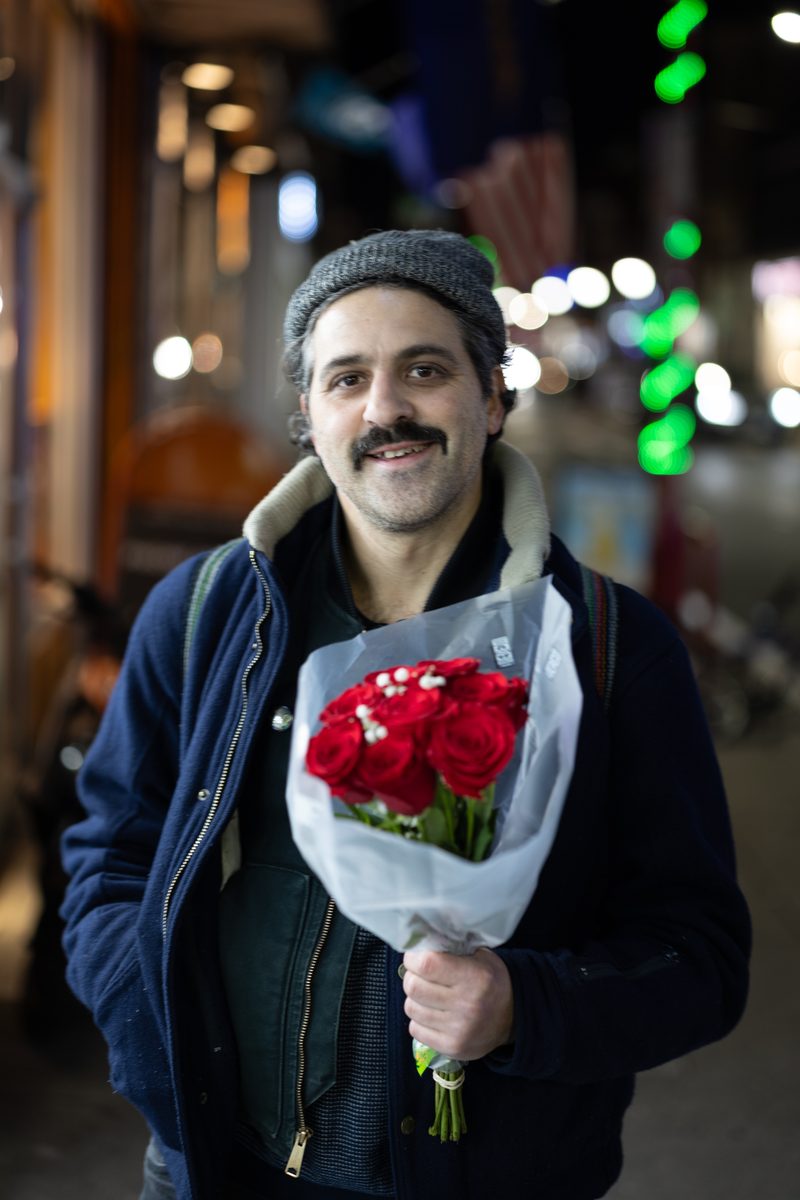 A smiling man with a mustache and gray beanie holds a bouquet of red roses on South Street at night.