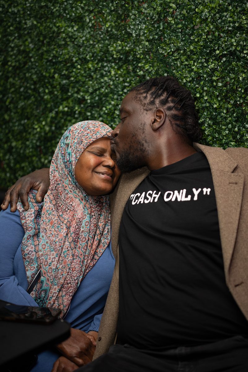 A man in a "Cash Only" t-shirt kisses the forehead of a woman in a floral hijab, seated together against a greenery wall at Room 148 Lounge on South Street.