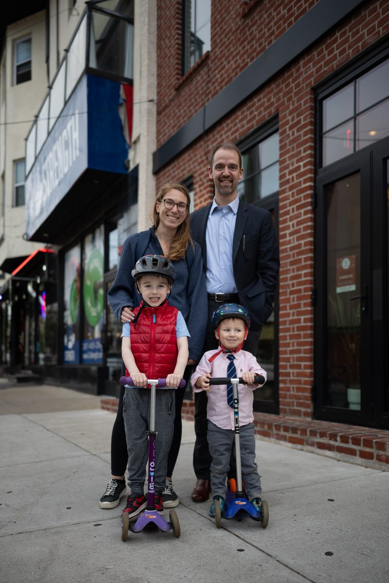 A family of four on a South Street sidewalk, the two young sons in helmets on scooters, Dad in a button-up shirt and blazer.