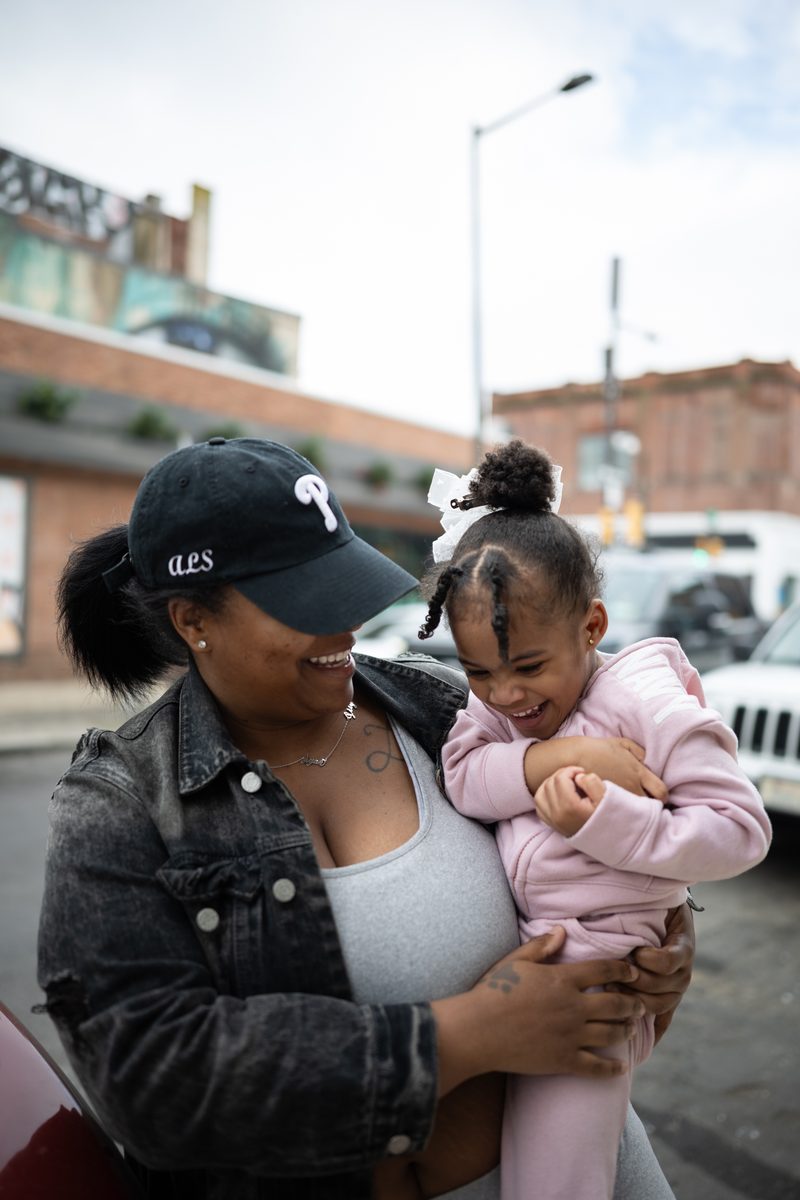 A mother in a Phillies cap laughs while holding her daughter in pink, whose hair is tied in bows, on South Street.