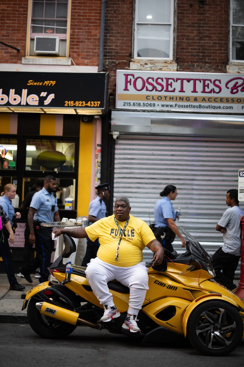 A man in a yellow "Funcle" T-shirt sits on a yellow three-wheeled motorcycle outside Ishkabibble's cheesesteak shop on South Street, a group of police officers walking behind him.